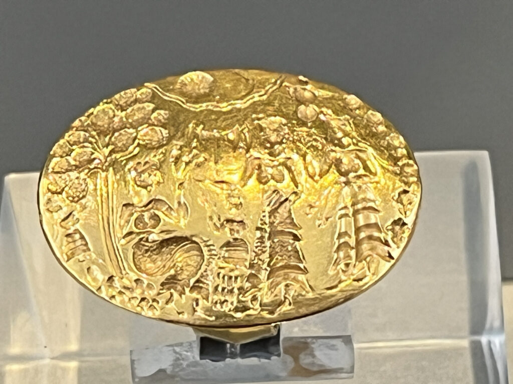 A gold ring featuring two women offer a sacrifice of lilies and poppies to a goddess sitting under a tree.